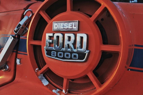 Close-up of an orange Ford 8000 diesel tractor badge featuring a bold, metallic logo with visible signs of wear and some areas of chipped paint.