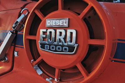 Close-up of an orange Ford 8000 diesel tractor badge featuring a bold, metallic logo with visible signs of wear and some areas of chipped paint.