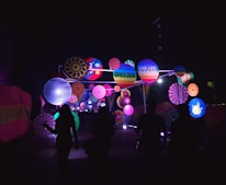 A vibrant outdoor installation features an array of colorful circular patterns and decorations illuminated against the dark night sky. Silhouettes of people are walking towards the brightly lit display, which includes playful designs and text such as 'Breezer' and 'Live Life in Colour.'