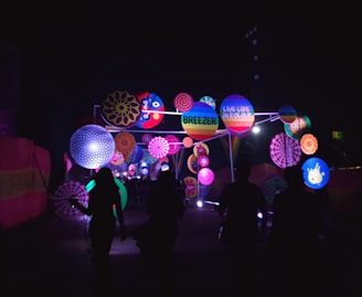 A vibrant outdoor installation features an array of colorful circular patterns and decorations illuminated against the dark night sky. Silhouettes of people are walking towards the brightly lit display, which includes playful designs and text such as 'Breezer' and 'Live Life in Colour.'