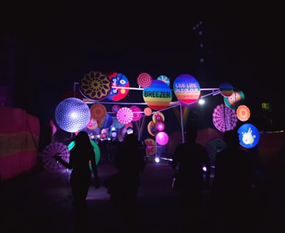 A vibrant outdoor installation features an array of colorful circular patterns and decorations illuminated against the dark night sky. Silhouettes of people are walking towards the brightly lit display, which includes playful designs and text such as 'Breezer' and 'Live Life in Colour.'