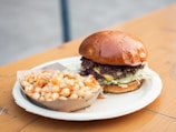 A delicious looking burger with a shiny bun, layers of lettuce, pickles, and a juicy beef patty covered in melted cheese sits on a paper plate. Next to it is a side of chickpea salad in a small cardboard tray, featuring diced vegetables and a hint of dressing. The setting appears to be on an outdoor wooden table, providing a casual dining feel.