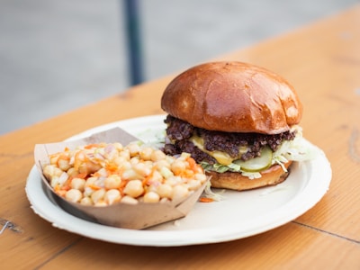 A delicious looking burger with a shiny bun, layers of lettuce, pickles, and a juicy beef patty covered in melted cheese sits on a paper plate. Next to it is a side of chickpea salad in a small cardboard tray, featuring diced vegetables and a hint of dressing. The setting appears to be on an outdoor wooden table, providing a casual dining feel.