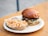 A delicious looking burger with a shiny bun, layers of lettuce, pickles, and a juicy beef patty covered in melted cheese sits on a paper plate. Next to it is a side of chickpea salad in a small cardboard tray, featuring diced vegetables and a hint of dressing. The setting appears to be on an outdoor wooden table, providing a casual dining feel.