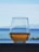 A glass filled with a light brown beverage is placed on a ledge overlooking a tranquil seascape. The background features a clear blue sky with light clouds and calm ocean waters, with faint outlines of distant mountains.