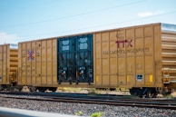 A freight train boxcar with a yellow exterior is parked on a set of railroad tracks. The side of the boxcar has the letters 'TTX' and the phrase 'RAILCAR POOLING EXPERTS' prominently displayed. The boxcar has instructions printed on the doors, and various mechanical details like wheels and coupling mechanisms are visible.