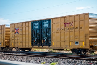 A freight train boxcar with a yellow exterior is parked on a set of railroad tracks. The side of the boxcar has the letters 'TTX' and the phrase 'RAILCAR POOLING EXPERTS' prominently displayed. The boxcar has instructions printed on the doors, and various mechanical details like wheels and coupling mechanisms are visible.