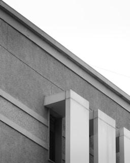 A section of a modern building with a minimalist design, featuring geometric patterns and clean lines. The facade is composed of textured concrete and smooth paneling. The perspective includes sharp angles and a high contrast between light and shadow.