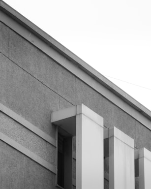 A section of a modern building with a minimalist design, featuring geometric patterns and clean lines. The facade is composed of textured concrete and smooth paneling. The perspective includes sharp angles and a high contrast between light and shadow.