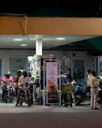 At a gas station, several individuals on motorbikes are gathered around fuel pumps under artificial lighting. Most people are wearing helmets and some have masks. The station features posters and signs in the background, with a prominent NFC payment poster visible.