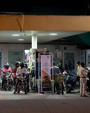 At a gas station, several individuals on motorbikes are gathered around fuel pumps under artificial lighting. Most people are wearing helmets and some have masks. The station features posters and signs in the background, with a prominent NFC payment poster visible.