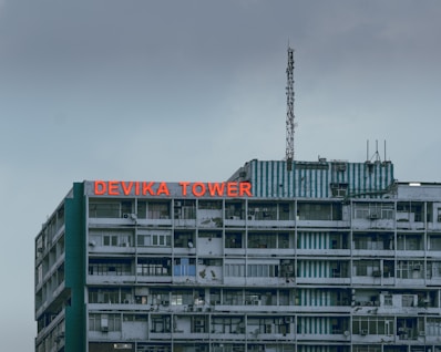 A multistory building with a weathered facade and a large red sign reading 'DEVIKA TOWER' on the top floor. The structure features various windows and balconies, some with visible air conditioning units. An antenna is mounted on the roof along with some other electronic equipment. The sky appears overcast, adding a muted tone to the overall scene.