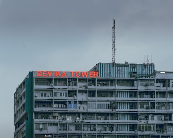 A multistory building with a weathered facade and a large red sign reading 'DEVIKA TOWER' on the top floor. The structure features various windows and balconies, some with visible air conditioning units. An antenna is mounted on the roof along with some other electronic equipment. The sky appears overcast, adding a muted tone to the overall scene.