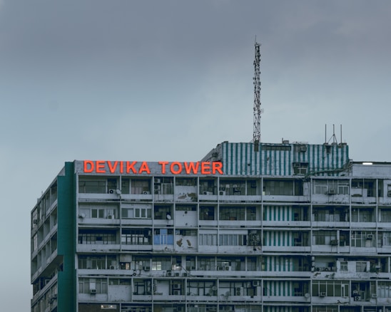 A multistory building with a weathered facade and a large red sign reading 'DEVIKA TOWER' on the top floor. The structure features various windows and balconies, some with visible air conditioning units. An antenna is mounted on the roof along with some other electronic equipment. The sky appears overcast, adding a muted tone to the overall scene.