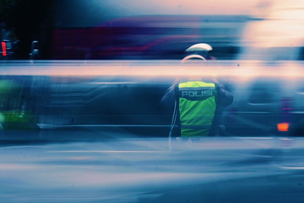 A person wearing a fluorescent yellow vest with 'Polisi' written on the back, possibly a traffic officer, stands amidst a motion-blurred background of vehicles. The scene conveys a sense of speed and movement.