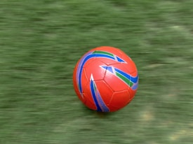 A vibrant red soccer ball with colorful blue and green geometric patterns is moving across a lush, green grass field. The motion blur suggests the ball is rolling quickly.