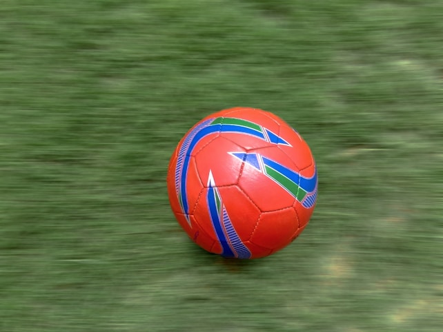 A vibrant red soccer ball with colorful blue and green geometric patterns is moving across a lush, green grass field. The motion blur suggests the ball is rolling quickly.