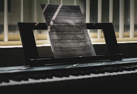 A sheet of music is propped up on an electronic keyboard, illuminated by soft light filtering through vertical blinds. Shadows and light play across the keys and create a serene, contemplative atmosphere.