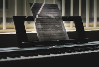 A sheet of music is propped up on an electronic keyboard, illuminated by soft light filtering through vertical blinds. Shadows and light play across the keys and create a serene, contemplative atmosphere.