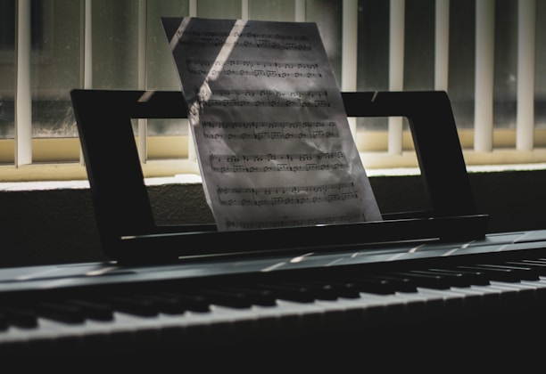 A sheet of music is propped up on an electronic keyboard, illuminated by soft light filtering through vertical blinds. Shadows and light play across the keys and create a serene, contemplative atmosphere.