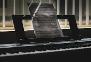 A sheet of music is propped up on an electronic keyboard, illuminated by soft light filtering through vertical blinds. Shadows and light play across the keys and create a serene, contemplative atmosphere.
