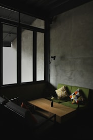 A dimly lit room with a minimalist design features large windows with dark frames that allow natural light to filter through. The space includes a green sofa adorned with patterned cushions, a simple wooden coffee table, and a black wall-mounted lamp. The overall ambiance is serene and understated.