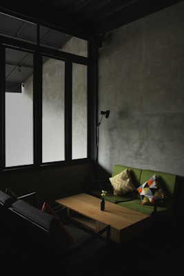A dimly lit room with a minimalist design features large windows with dark frames that allow natural light to filter through. The space includes a green sofa adorned with patterned cushions, a simple wooden coffee table, and a black wall-mounted lamp. The overall ambiance is serene and understated.