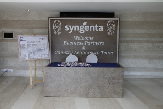A welcome area with a large sign featuring the logo 'syngenta' and a message for business partners and country leadership team is displayed on a wall. There is a seating plan on a wooden easel to the left, and a table with wrapped items in front of the sign.