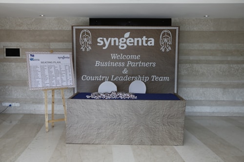 A welcome area with a large sign featuring the logo 'syngenta' and a message for business partners and country leadership team is displayed on a wall. There is a seating plan on a wooden easel to the left, and a table with wrapped items in front of the sign.