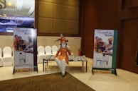 A conference hall with a display featuring two promotional standees showcasing a family in traditional attire, highlighting a product related to agriculture. There is a statue of a man seated with a child on his shoulders and a patterned table. The lighting is warm and the decor includes elegant paneling and chandeliers.