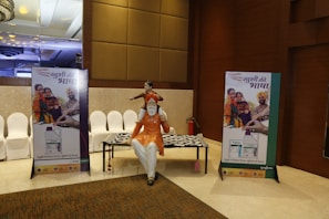 A conference hall with a display featuring two promotional standees showcasing a family in traditional attire, highlighting a product related to agriculture. There is a statue of a man seated with a child on his shoulders and a patterned table. The lighting is warm and the decor includes elegant paneling and chandeliers.