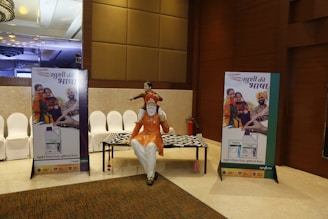 A conference hall with a display featuring two promotional standees showcasing a family in traditional attire, highlighting a product related to agriculture. There is a statue of a man seated with a child on his shoulders and a patterned table. The lighting is warm and the decor includes elegant paneling and chandeliers.