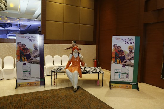 A conference hall with a display featuring two promotional standees showcasing a family in traditional attire, highlighting a product related to agriculture. There is a statue of a man seated with a child on his shoulders and a patterned table. The lighting is warm and the decor includes elegant paneling and chandeliers.
