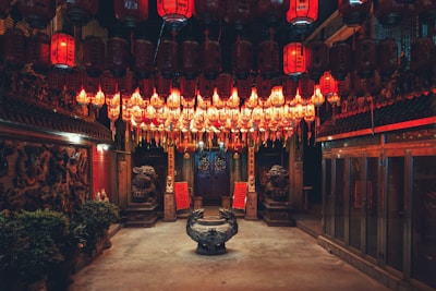 A richly decorated interior space is filled with rows of hanging red lanterns casting a warm glow. Stone statues of mythological creatures guard a central altar, surrounded by intricate carvings and traditional architecture. The atmosphere is serene and reverent.