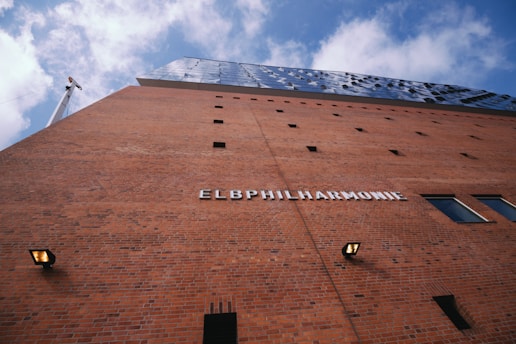 A large modern building with a brick facade and a metallic top section set against a partly cloudy sky. The facade features the word 'ELBPHILHARMONIE' in large white letters.