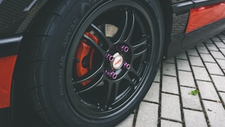A close-up view of a car's wheel showcasing a black alloy rim with purple lug nuts and a visible red brake caliper. The car tire is mounted on a pavement made of rectangular concrete bricks. The branding on the wheel and tire is prominent.