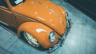 A vintage orange car with a classic design features prominently in a showroom or garage setting. The car's surface is glossy with visible reflections, and it has prominent rounded fenders and chrome detailing.