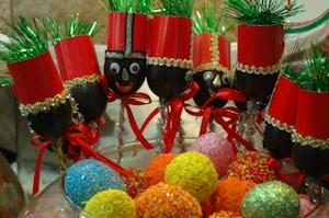 A group of festive crafts are made from wine glasses decorated as nutcracker soldiers. Each glass is topped with red paper hats and green tinsel, and the stems are adorned with gold and silver ribbons, red bows, and googly eyes. They are displayed in a decorative manner alongside an assortment of colorful, textured balls resembling candies.
