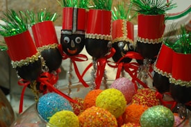 A group of festive crafts are made from wine glasses decorated as nutcracker soldiers. Each glass is topped with red paper hats and green tinsel, and the stems are adorned with gold and silver ribbons, red bows, and googly eyes. They are displayed in a decorative manner alongside an assortment of colorful, textured balls resembling candies.