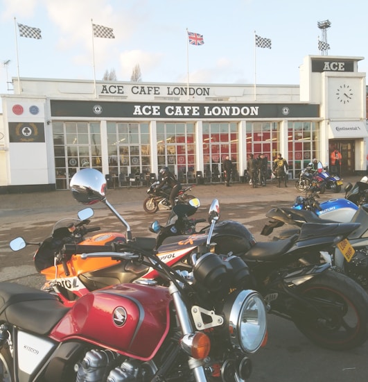 A lineup of motorcycles is parked outside a retro-style caf&eacute;. The building has a prominent sign reading 'ACE CAFE LONDON' above the entrance. Several people are gathered near the entrance, some clad in motorcycle gear. Flags, including a British flag and checkered racing flags, are displayed on poles atop the building.