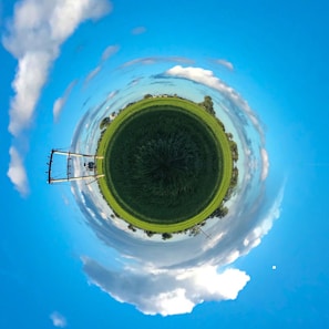A panoramic, spherical view featuring a grassy field at the center, surrounded by a 360-degree horizon with vibrant blue skies and scattered clouds. An electricity pole is visible on the left side, adding an element of infrastructure amidst the natural scenery.
