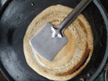 A round, golden-brown crepe or dosa is cooking on a black frying pan. A flat, stainless steel spatula is placed on top of the crepe.