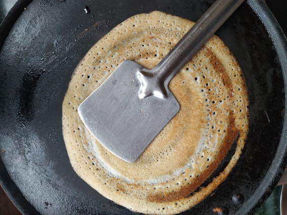 A round, golden-brown crepe or dosa is cooking on a black frying pan. A flat, stainless steel spatula is placed on top of the crepe.