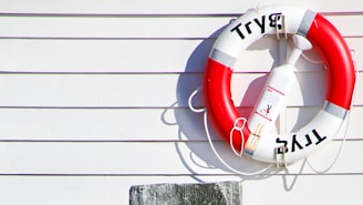 A lifebuoy with red and white sections is mounted on a light-colored horizontal wooden siding. The lifebuoy has the word 'Tryg' on it, and a small buoy is attached to it with a rope. The shadow of the lifebuoy is cast onto the wall by sunlight.