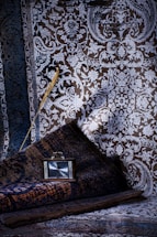 An intricate, richly patterned fabric with detailed floral and geometric designs in shades of blue and brown dominates the background. A rolled rug rests at an angle, featuring similar elaborate patterns. A small, vintage-style clock with a dark wooden frame rests on the rug, displaying the time. An elegant, gold-tinted feather lies across the fabric, adding a touch of contrast and texture.