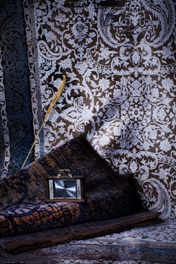 An intricate, richly patterned fabric with detailed floral and geometric designs in shades of blue and brown dominates the background. A rolled rug rests at an angle, featuring similar elaborate patterns. A small, vintage-style clock with a dark wooden frame rests on the rug, displaying the time. An elegant, gold-tinted feather lies across the fabric, adding a touch of contrast and texture.