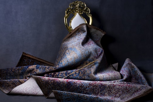 A richly textured, ornate rug with a complex pattern is draped over a decorative chair with a gold-toned frame and an oval white backrest. The rug displays a mix of blue, red, and beige colors with an intricate design. The setting is dimly lit, creating a dramatic and elegant atmosphere.
