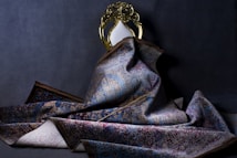 A richly textured, ornate rug with a complex pattern is draped over a decorative chair with a gold-toned frame and an oval white backrest. The rug displays a mix of blue, red, and beige colors with an intricate design. The setting is dimly lit, creating a dramatic and elegant atmosphere.