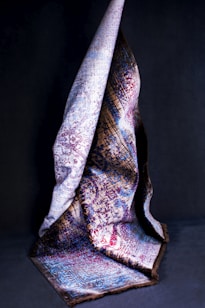 A richly textured, multicolored fabric is draped artistically against a dark background. The fabric features intricate patterns in hues of blue, red, and brown, with portions of the cloth elegantly folded to showcase different sections of the design.