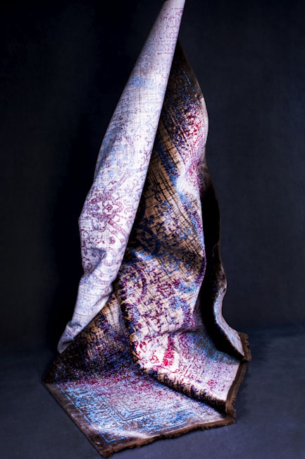 A richly textured, multicolored fabric is draped artistically against a dark background. The fabric features intricate patterns in hues of blue, red, and brown, with portions of the cloth elegantly folded to showcase different sections of the design.